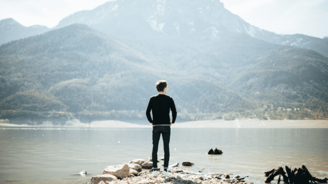 一名男子站在湖前的岩石上，背景是山，感到焦慮。