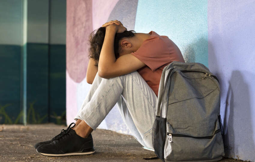A young man sitting on the ground with his backpack, exhibiting signs of anxiety.