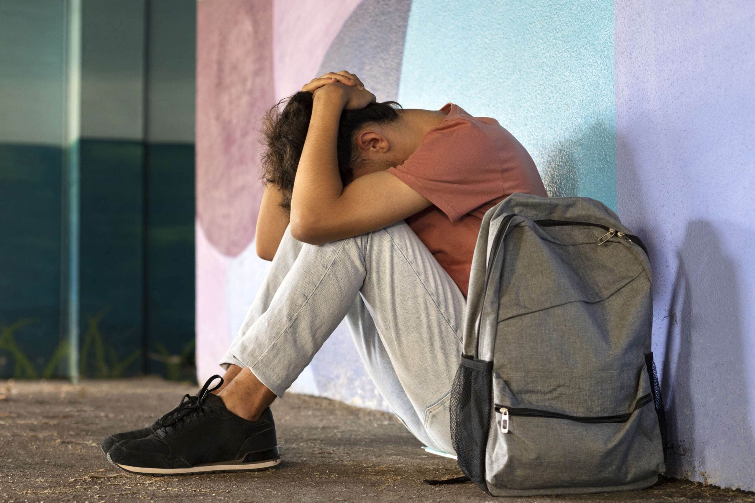A young man sitting on the ground with his backpack, exhibiting signs of anxiety.